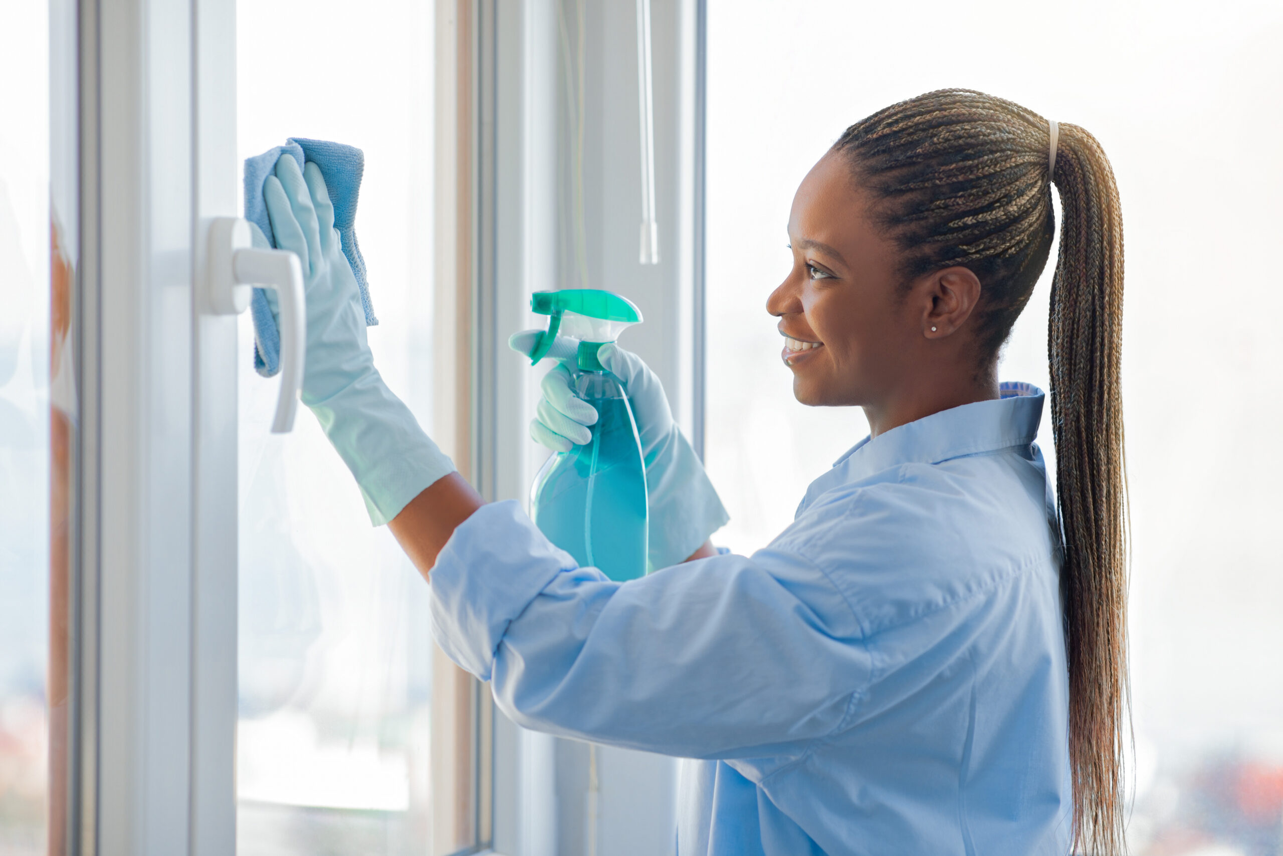 Cheerful happy pretty young black woman with long braids wearing homewear housewife cleaning windows at home and smiling, enjoying chores, using duster and spray, side view